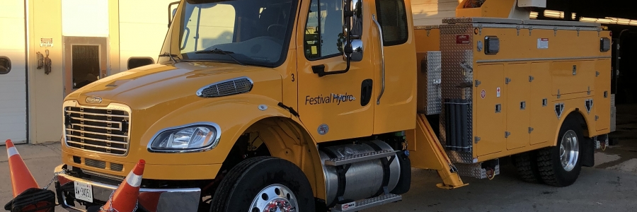 a yellow festival hydro bucket truck is parked in front of a yellow brick building with an open bay door.