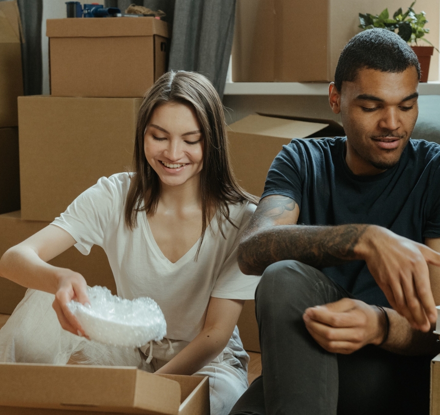 start a man in a blue t-shirt and a woman in a white t-shirt sit among cardboard boxes. The woman is unpacking a white bowl wrapped in bubble wrap.