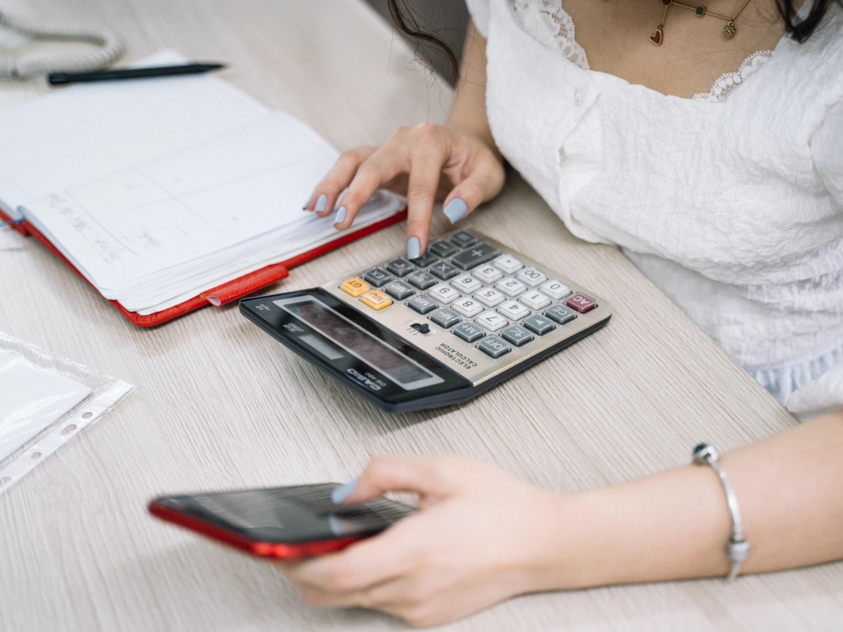 a woman holding a red iphone, typing on a calculatore with a red notebook laying on the desk to her right.