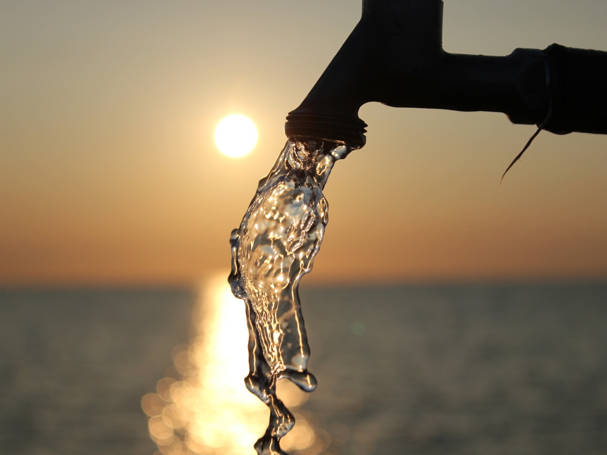 A running faucet is pictured with a sunsetting over a lake in the background.