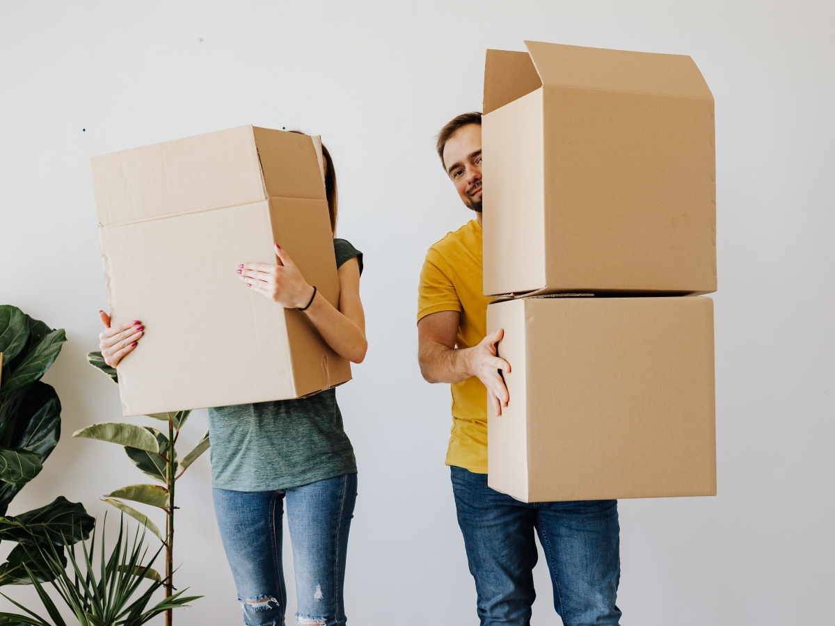 two peope in blue jeans are holding cardboard boxes and standing in fromt of a white wall with some plants on the left.